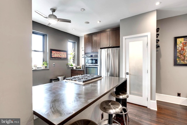 kitchen with dark brown cabinetry, ceiling fan, dark wood-type flooring, a kitchen bar, and appliances with stainless steel finishes