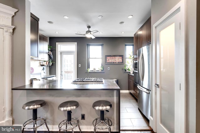 kitchen with kitchen peninsula, dark brown cabinetry, ceiling fan, light tile patterned floors, and stainless steel refrigerator