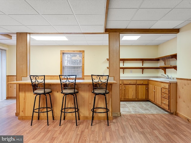 kitchen with kitchen peninsula, a drop ceiling, a breakfast bar, wooden walls, and light hardwood / wood-style floors