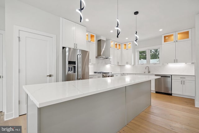 kitchen with light wood-type flooring, wall chimney exhaust hood, stainless steel appliances, white cabinetry, and a kitchen island