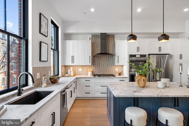 kitchen with wall chimney range hood, appliances with stainless steel finishes, sink, and light stone counters