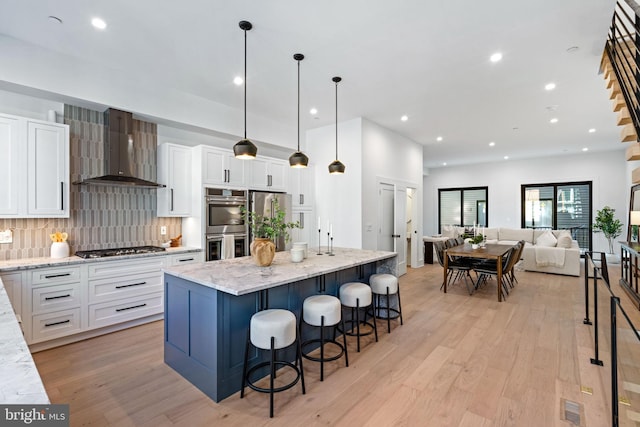 kitchen featuring light hardwood / wood-style floors, an island with sink, wall chimney range hood, white cabinetry, and decorative light fixtures