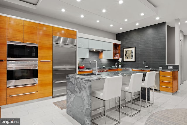 kitchen with a kitchen breakfast bar, built in appliances, dark stone counters, a spacious island, and decorative backsplash