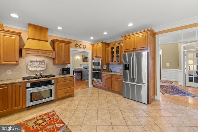 kitchen with backsplash, premium range hood, light stone counters, stainless steel appliances, and crown molding