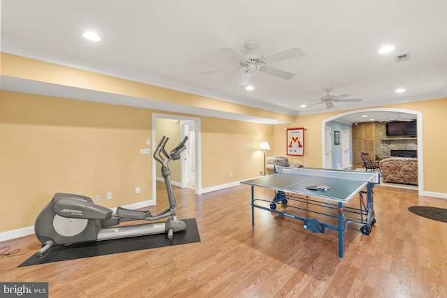 recreation room with a stone fireplace, ceiling fan, light hardwood / wood-style flooring, and crown molding