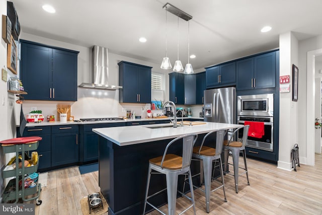 kitchen featuring a kitchen island with sink, sink, wall chimney exhaust hood, blue cabinetry, and appliances with stainless steel finishes