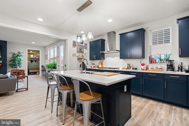 kitchen with blue cabinetry, light wood-type flooring, a kitchen island with sink, and wall chimney range hood