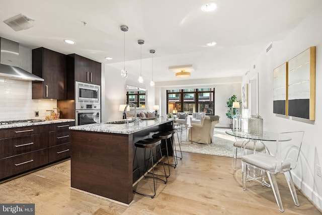 kitchen with appliances with stainless steel finishes, wall chimney exhaust hood, a center island with sink, light hardwood / wood-style flooring, and a breakfast bar area