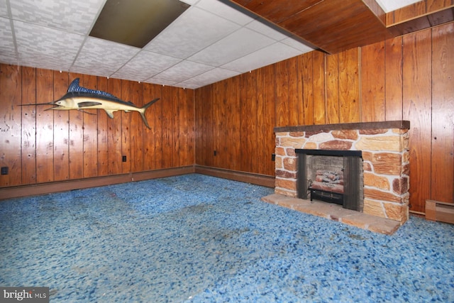 unfurnished living room with a drop ceiling, baseboard heating, wooden walls, a fireplace, and carpet