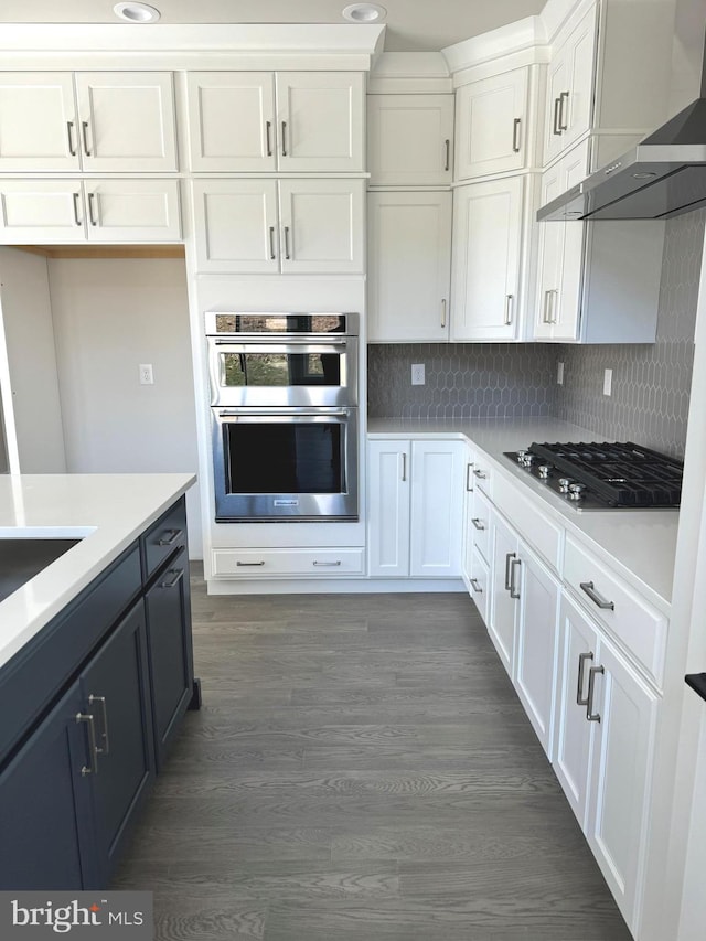 kitchen featuring tasteful backsplash, wall chimney range hood, dark hardwood / wood-style flooring, white cabinets, and double oven