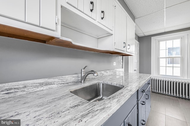 kitchen with radiator, a paneled ceiling, sink, light stone countertops, and white cabinetry