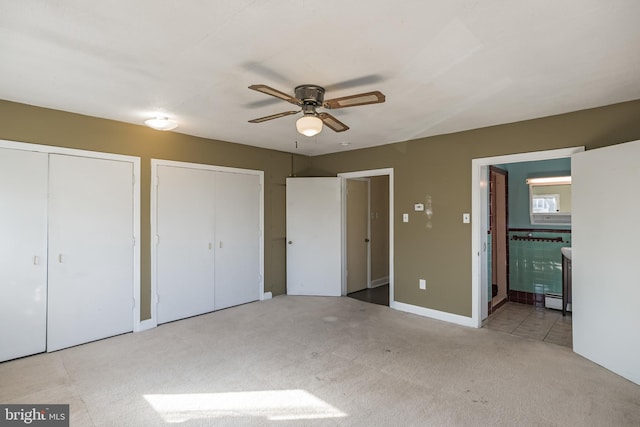 unfurnished bedroom with light carpet, baseboard heating, ceiling fan, connected bathroom, and multiple closets