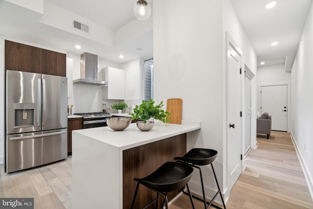 kitchen featuring a kitchen bar, wall chimney exhaust hood, light hardwood / wood-style flooring, and appliances with stainless steel finishes