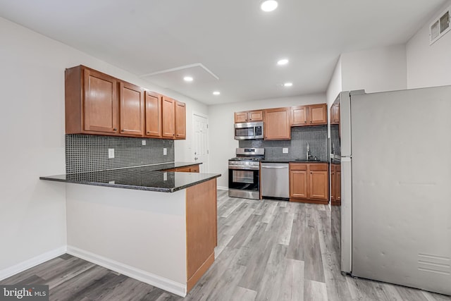 kitchen with kitchen peninsula, appliances with stainless steel finishes, backsplash, dark stone counters, and light hardwood / wood-style flooring