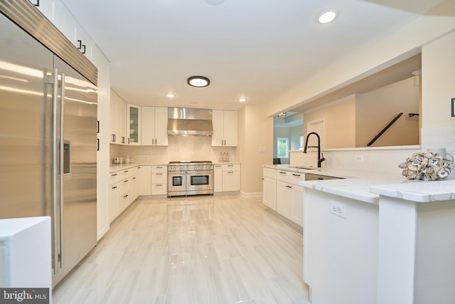 kitchen featuring white cabinetry, high end appliances, wall chimney exhaust hood, kitchen peninsula, and backsplash