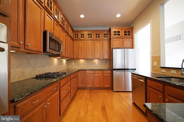 kitchen with appliances with stainless steel finishes, light wood-type flooring, backsplash, dark stone counters, and sink