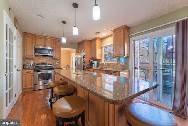 kitchen featuring light wood-type flooring, a textured ceiling, stainless steel appliances, decorative light fixtures, and dark stone countertops