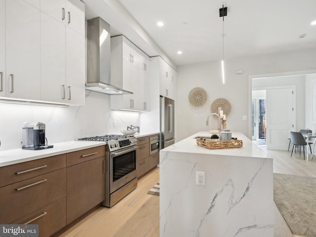 kitchen with wall chimney exhaust hood, premium appliances, decorative light fixtures, white cabinets, and light wood-type flooring