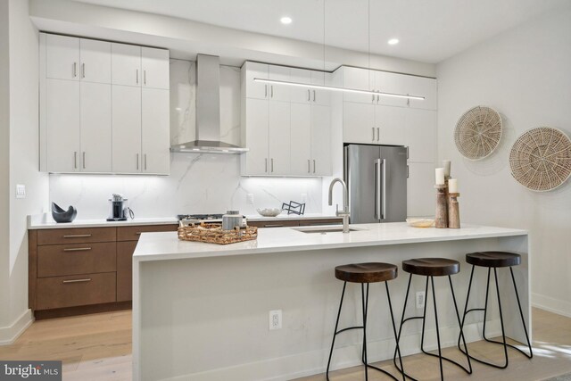 kitchen featuring white cabinets, high end refrigerator, a center island with sink, and wall chimney exhaust hood