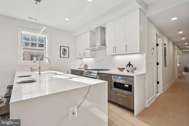 kitchen featuring light stone countertops, stainless steel appliances, wall chimney range hood, decorative light fixtures, and white cabinets