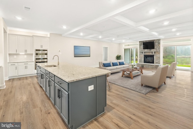 kitchen featuring a stone fireplace, light wood-type flooring, an island with sink, light stone counters, and white cabinetry