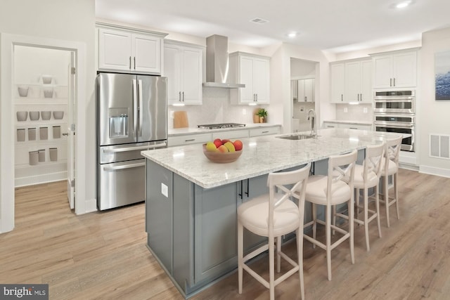 kitchen with light stone countertops, appliances with stainless steel finishes, sink, wall chimney range hood, and a center island with sink