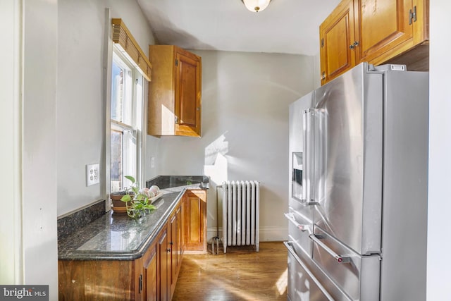 kitchen with dark wood-style floors, high end fridge, brown cabinets, dark stone counters, and radiator heating unit