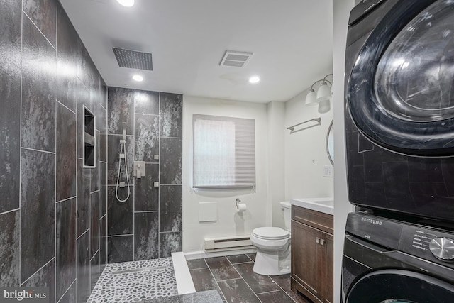 bathroom with vanity, baseboard heating, tiled shower, stacked washer and clothes dryer, and toilet