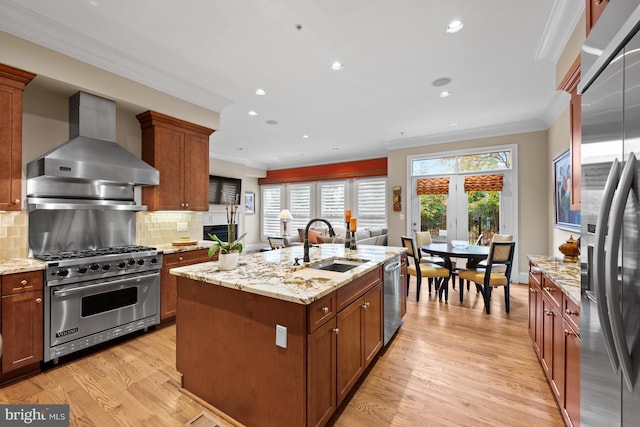 kitchen with stainless steel appliances, a kitchen island with sink, sink, wall chimney range hood, and light hardwood / wood-style flooring