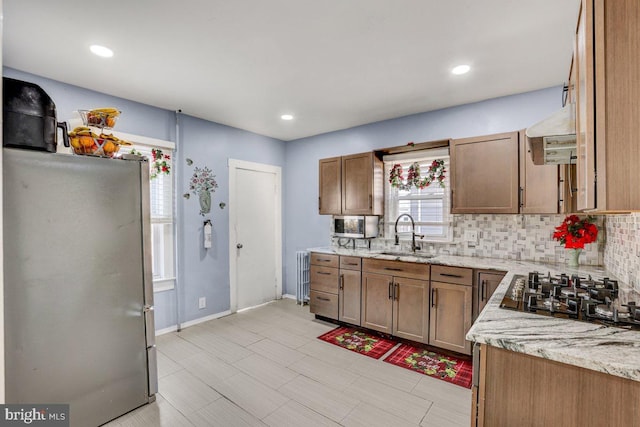 kitchen with exhaust hood, black gas stovetop, sink, stainless steel fridge, and light stone countertops