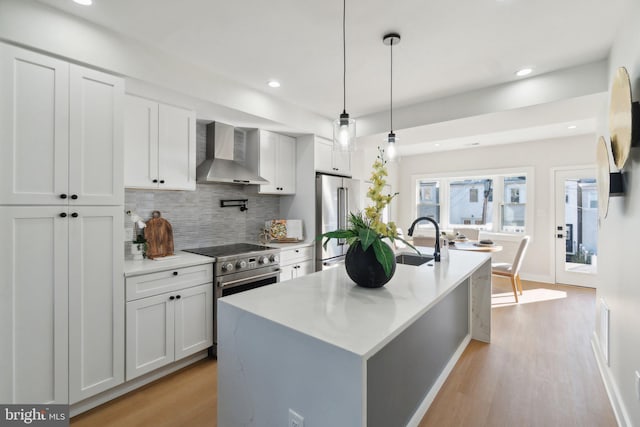 kitchen with wall chimney exhaust hood, stainless steel appliances, hanging light fixtures, an island with sink, and white cabinets