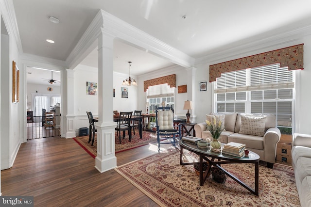 living room featuring crown molding, dark wood-type flooring, ceiling fan with notable chandelier, and ornate columns