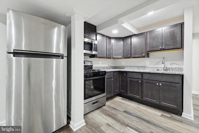 kitchen with appliances with stainless steel finishes, light wood-type flooring, light stone counters, dark brown cabinets, and sink