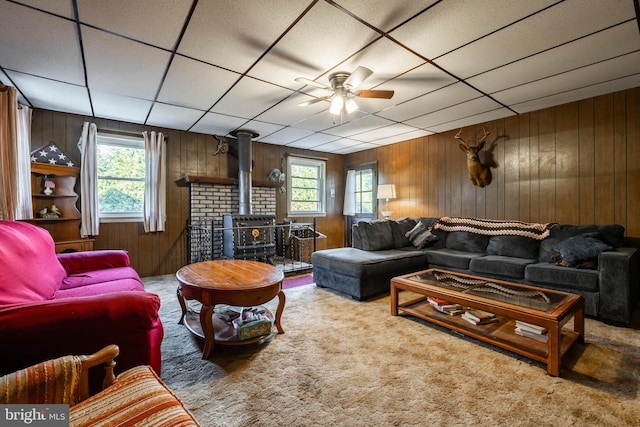 carpeted living room with ceiling fan, wood walls, a drop ceiling, and a wood stove