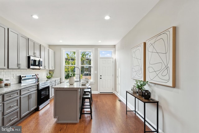 kitchen with gray cabinetry, light stone counters, a kitchen bar, a kitchen island with sink, and appliances with stainless steel finishes