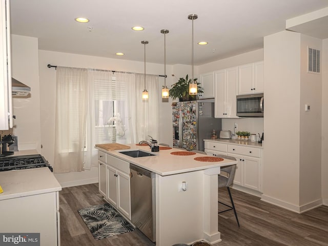 kitchen with wall chimney range hood, decorative light fixtures, a kitchen island with sink, white cabinets, and appliances with stainless steel finishes