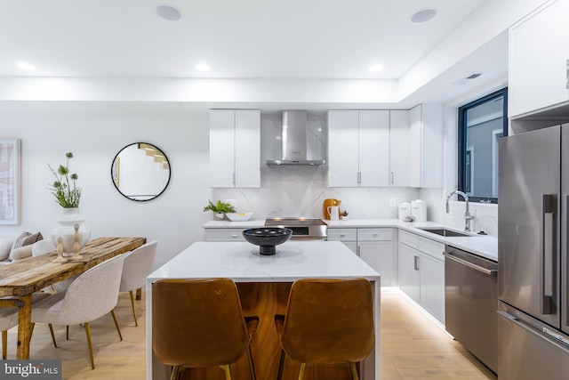 kitchen with wall chimney exhaust hood, light wood-type flooring, appliances with stainless steel finishes, a kitchen island, and white cabinetry