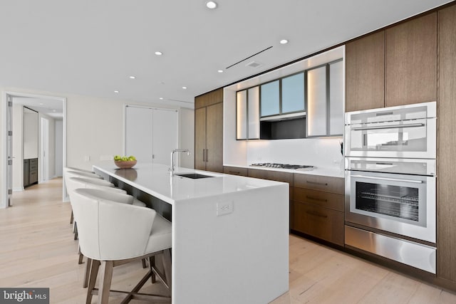 kitchen with a center island with sink, sink, light hardwood / wood-style flooring, stainless steel double oven, and gas cooktop