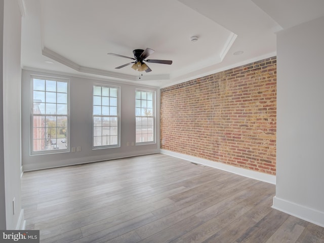 empty room with light hardwood / wood-style flooring, a raised ceiling, ceiling fan, and brick wall