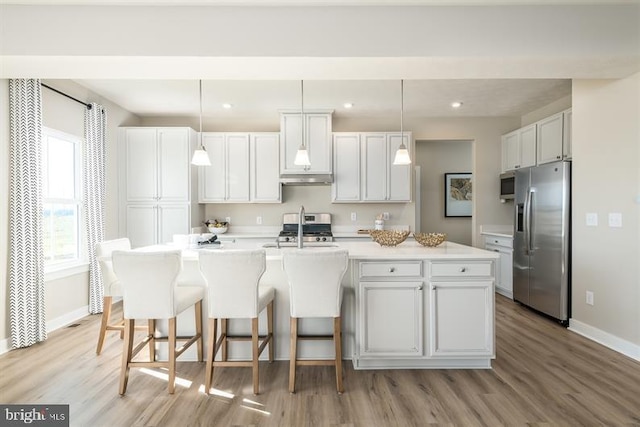 kitchen with appliances with stainless steel finishes, decorative light fixtures, a center island with sink, white cabinets, and light wood-type flooring