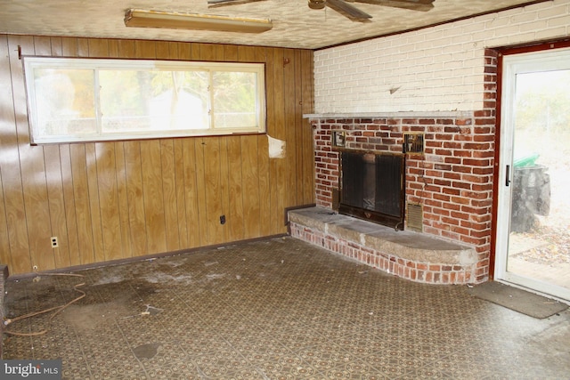 unfurnished living room with a brick fireplace, ceiling fan, wooden walls, and brick wall