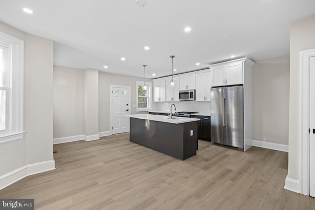 kitchen with appliances with stainless steel finishes, pendant lighting, light hardwood / wood-style floors, white cabinetry, and an island with sink