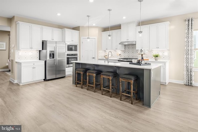 kitchen with appliances with stainless steel finishes, light wood-type flooring, a center island with sink, white cabinetry, and hanging light fixtures