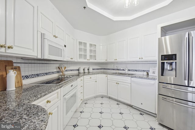 kitchen with a raised ceiling, backsplash, dark stone countertops, white appliances, and white cabinets