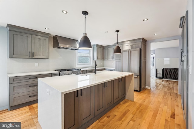 kitchen with high end fridge, wall chimney exhaust hood, pendant lighting, light hardwood / wood-style flooring, and an island with sink