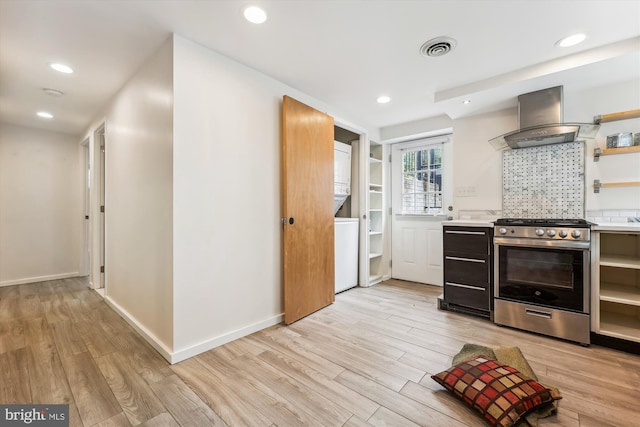 kitchen with wall chimney range hood, stacked washer and dryer, light hardwood / wood-style floors, and stainless steel range oven