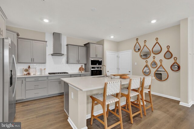 kitchen featuring wall chimney range hood, an island with sink, appliances with stainless steel finishes, dark hardwood / wood-style flooring, and a breakfast bar area