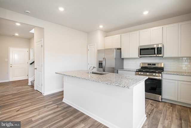 kitchen with sink, an island with sink, appliances with stainless steel finishes, light hardwood / wood-style floors, and light stone counters