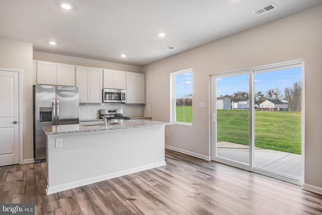 kitchen with light stone countertops, stainless steel appliances, white cabinetry, and an island with sink