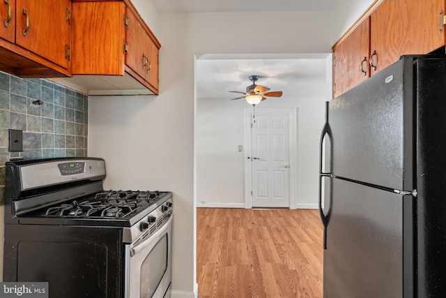 kitchen featuring ceiling fan, stainless steel gas range oven, decorative backsplash, black refrigerator, and light wood-type flooring